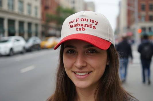 A woman sitting in a vehicle wearing a beige trucker cap with the text 'PUT IT ON MY HUSBAND'S TAB' embroidered on the front. Other similar caps are displayed in a row, showing different color options.