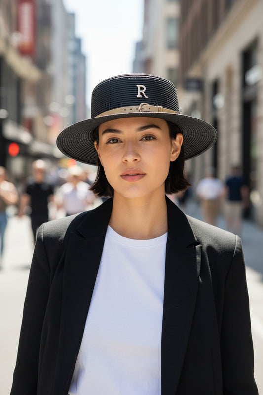 Black straw hat with a gold band and 'R' emblem held by a hand against a dark background